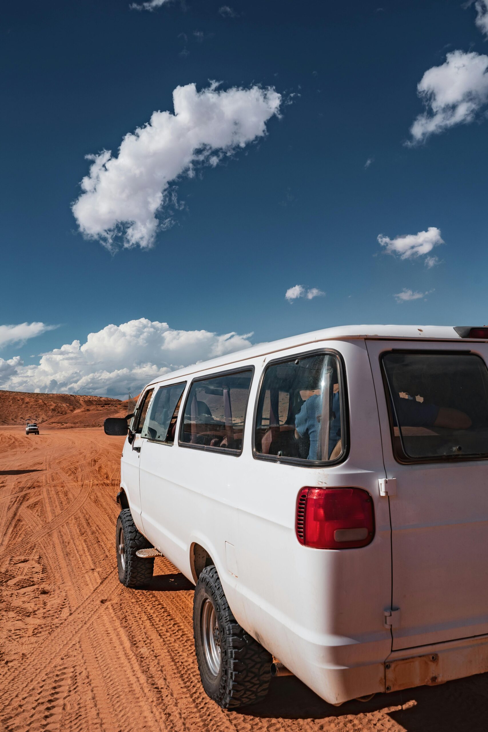 White van travels on a desert road under a clear blue sky with fluffy clouds.